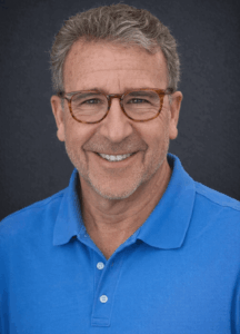 The image shows a person with short, gray hair wearing a blue shirt.