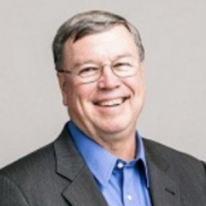 The image depicts an individual dressed in a dark suit with a blue shirt, wearing a polished, professional appearance.