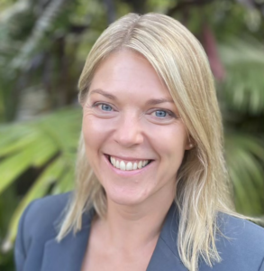 The image depicts a person with light blonde hair wearing a dark suit, surrounded by lush green foliage.